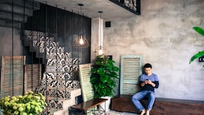 A man sitting on a wooden bench in a room with patterned tiles, a large plant, and hanging lights. - Memory Hostel (Da Nang)