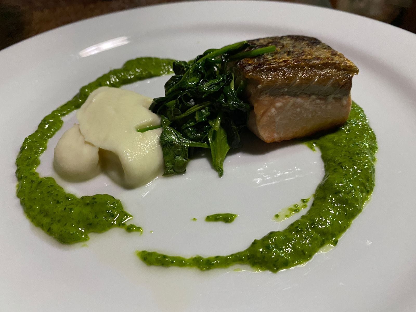 A plate with a piece of meat, greens, and a dollop of sauce.