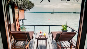 Una terraza de madera con dos sillas y una mesa, con vista a una masa de agua tranquila donde se divisa un velero a lo lejos.