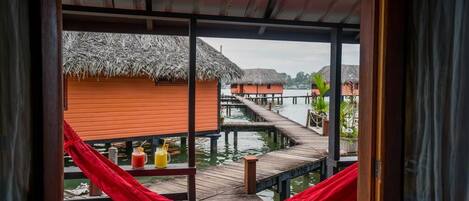 A view from a window overlooking a wooden dock with thatched-roof huts, a red hammock, and two drinks on the dock.