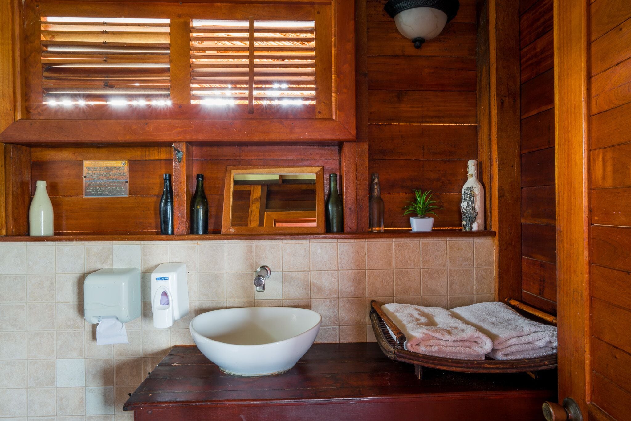 A wooden bathroom with a large vessel sink, a mirror, a towel rack, and a window with blinds.
