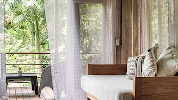 Ein Schlafzimmer mit einem hölzernen Bett, einem kleinen Tisch mit einer Pflanze und Blick auf üppiges Grün durch transparente Vorhänge.