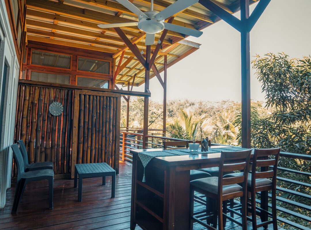 Un patio al aire libre con mesa y sillas de madera, un ventilador de techo y vistas del paisaje circundante.