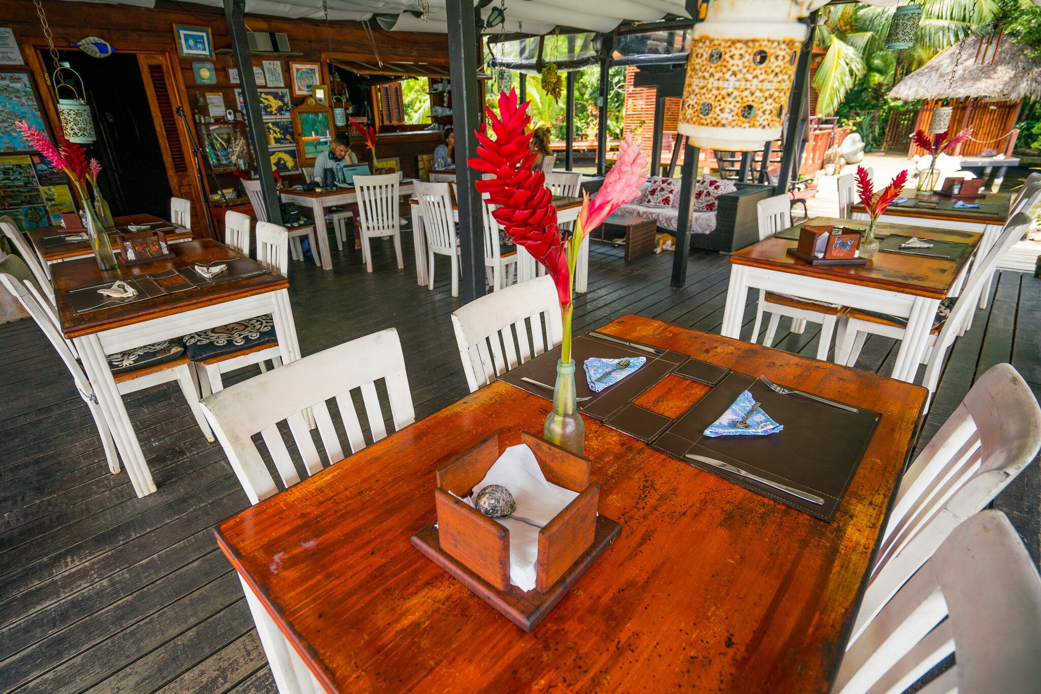 Un espace de restauration en plein air avec des tables en bois et des chaises blanches, entouré d’une terrasse en bois et couvert d’un toit.