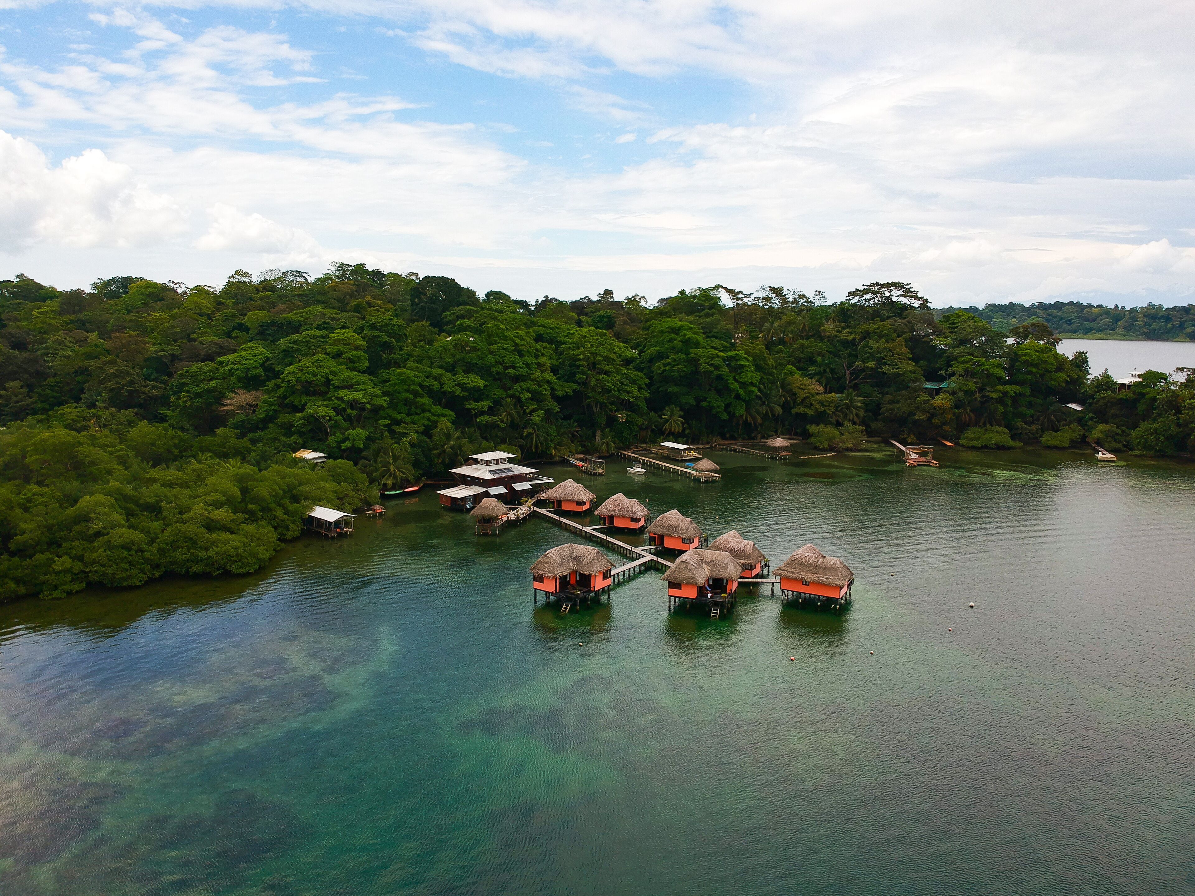 Vista aérea de un resort flotante con cabañas de techo de paja conectadas por pasarelas sobre aguas cristalinas, rodeado de densa selva.