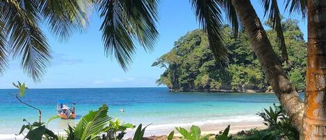 A tropical beach with clear blue water, a boat, and lush greenery.