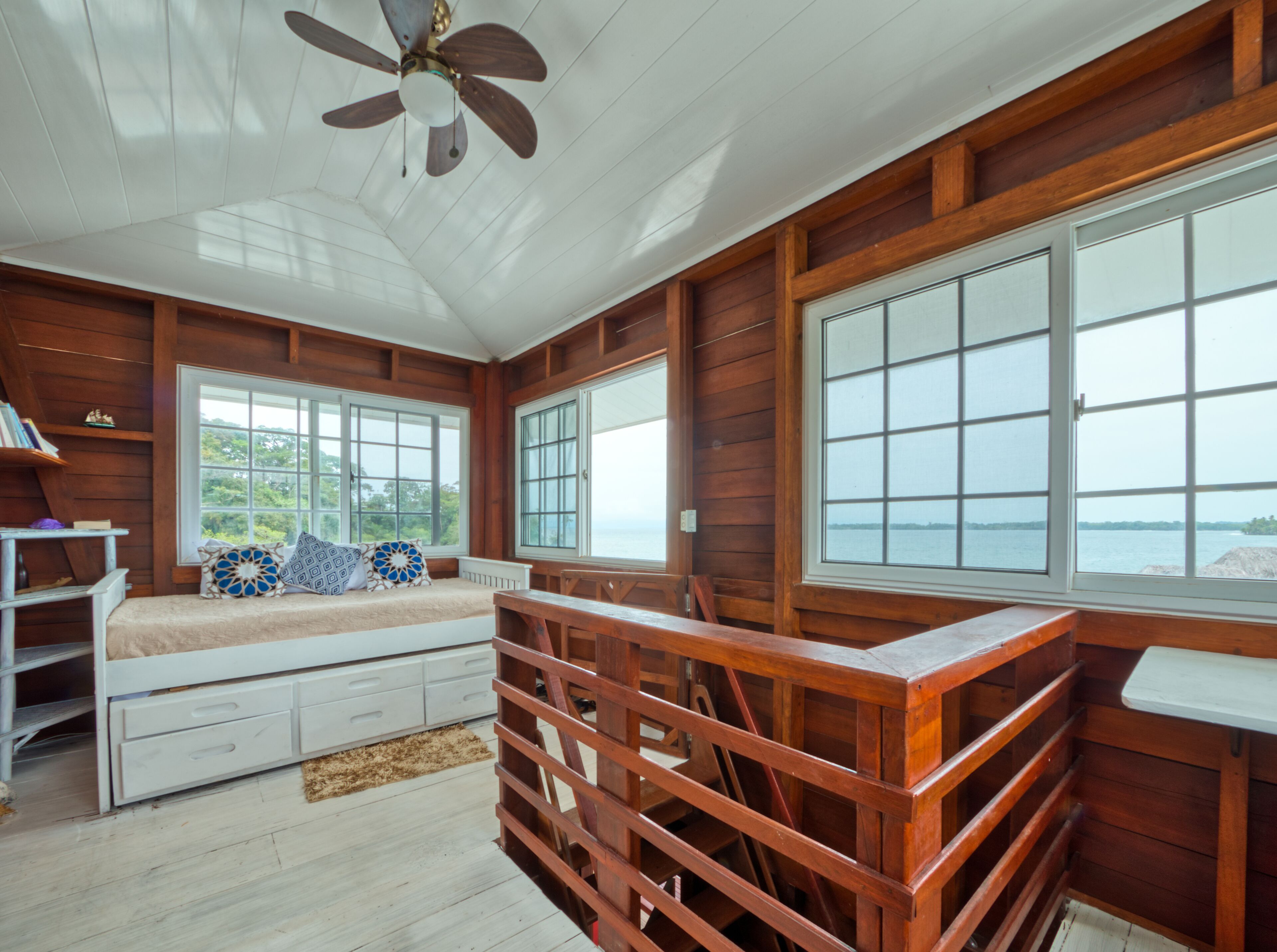 A wooden cabin with a bed, a desk, and large windows overlooking a body of water.