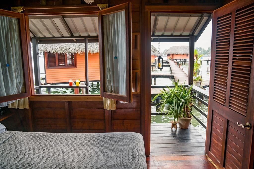 A wooden cabin with a balcony overlooking a water body, featuring a bed and a table with drinks.