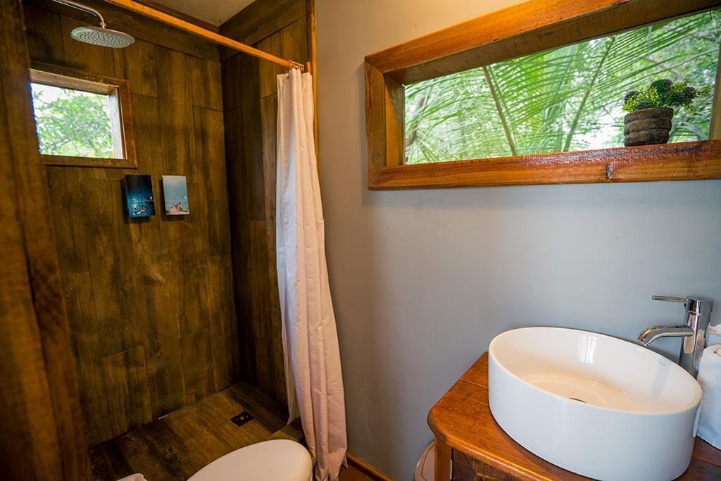 Un baño con cabina de ducha de madera, un lavamanos blanco y una ventana con vistas a follaje verde.