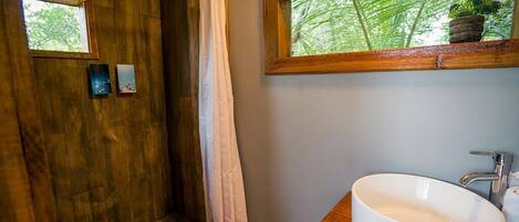 Un baño con cabina de ducha de madera, un lavamanos blanco y una ventana con vistas a follaje verde.