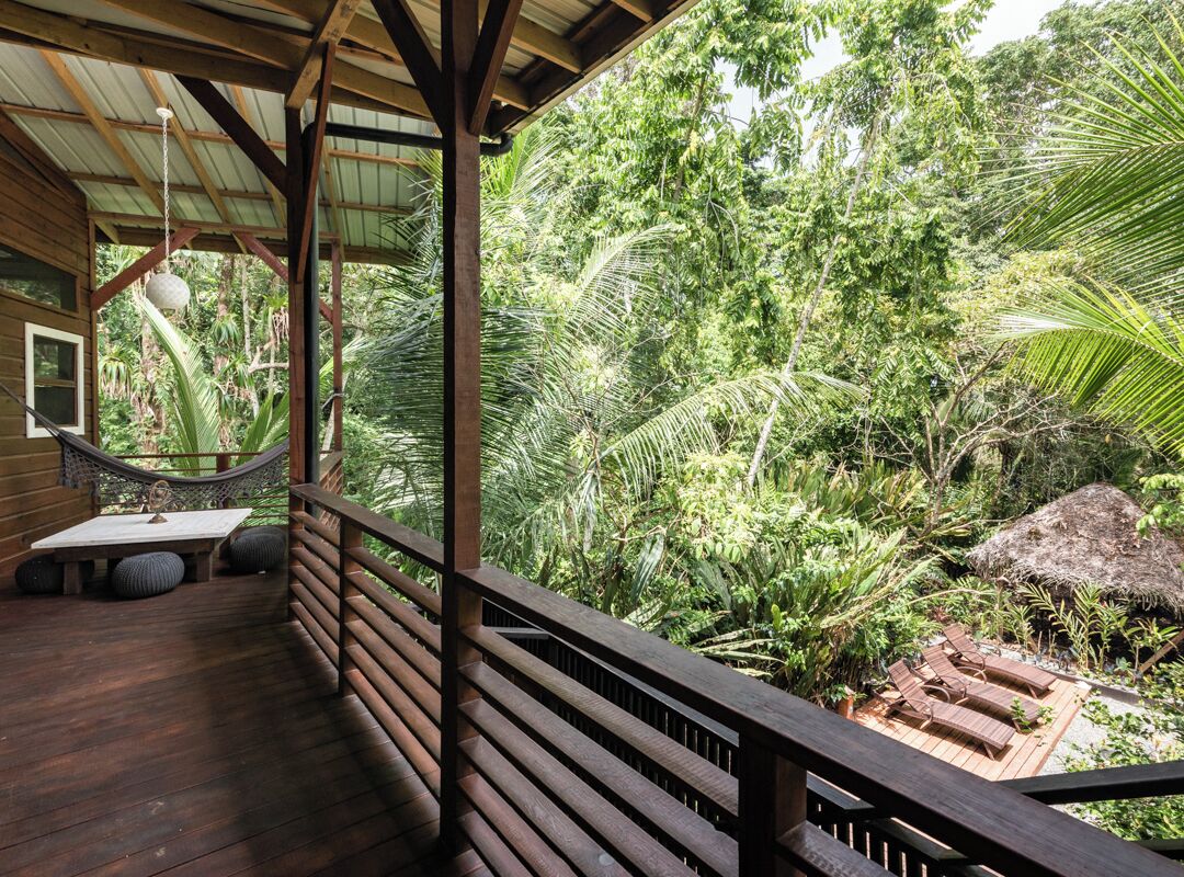 Un terrazzo in legno con un'amaca e un'area salotto con vista su un rigoglioso giardino tropicale.