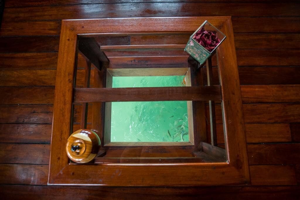 Una ventana de madera con vistas verdes, un pequeño objeto de madera y un recipiente de vidrio con elementos rojos.