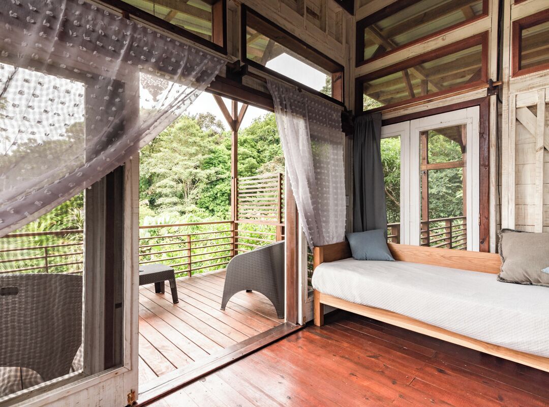 A wooden deck with a bed and a chair, a window with curtains, and a view of greenery outside.