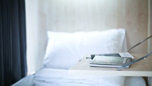 Ein ordentlich bezogenes Bett mit weißer Bettwäsche und Kissen, ein Nachttisch mit Buch und Stift sowie ein Fenster mit Vorhängen.