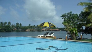 Ein Pool mit Liegestühlen und einem gelben Sonnenschirm, umgeben von Palmen und Blick auf ein Gewässer.