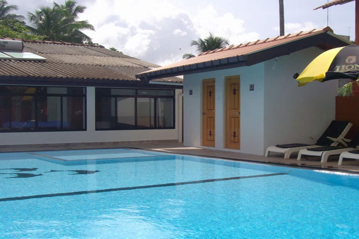 A swimming pool with lounge chairs and an umbrella, a building with large windows and doors, and a tiled roof.