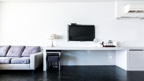 A modern living room with a white sofa, a small black stool, a flat-screen TV mounted on a white wall, and a white cabinet below. - Modena B&B (Taitung)