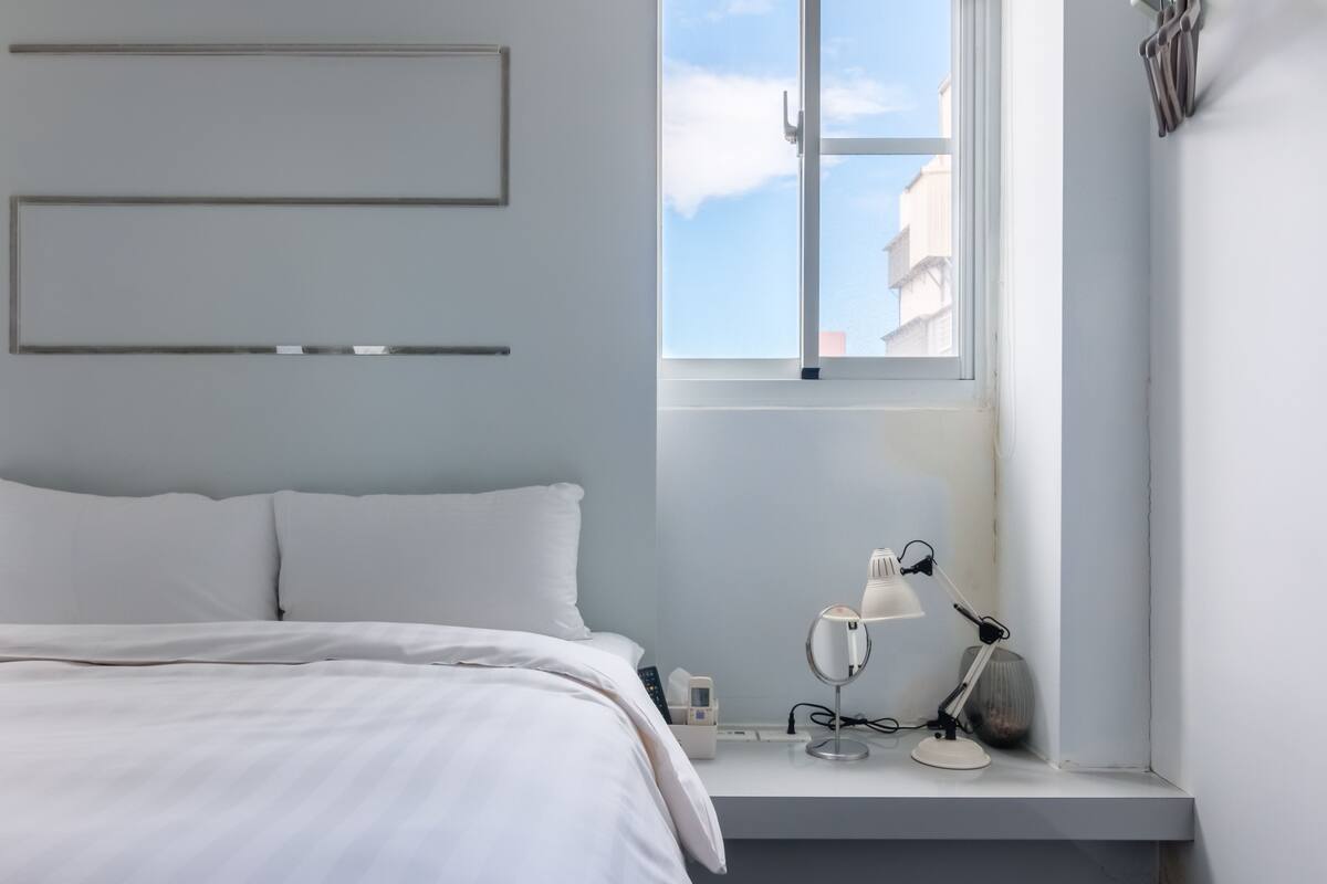 Een slaapkamer met een bed, een nachtkastje met een lamp, een raam met uitzicht op gebouwen en een wandplank.
