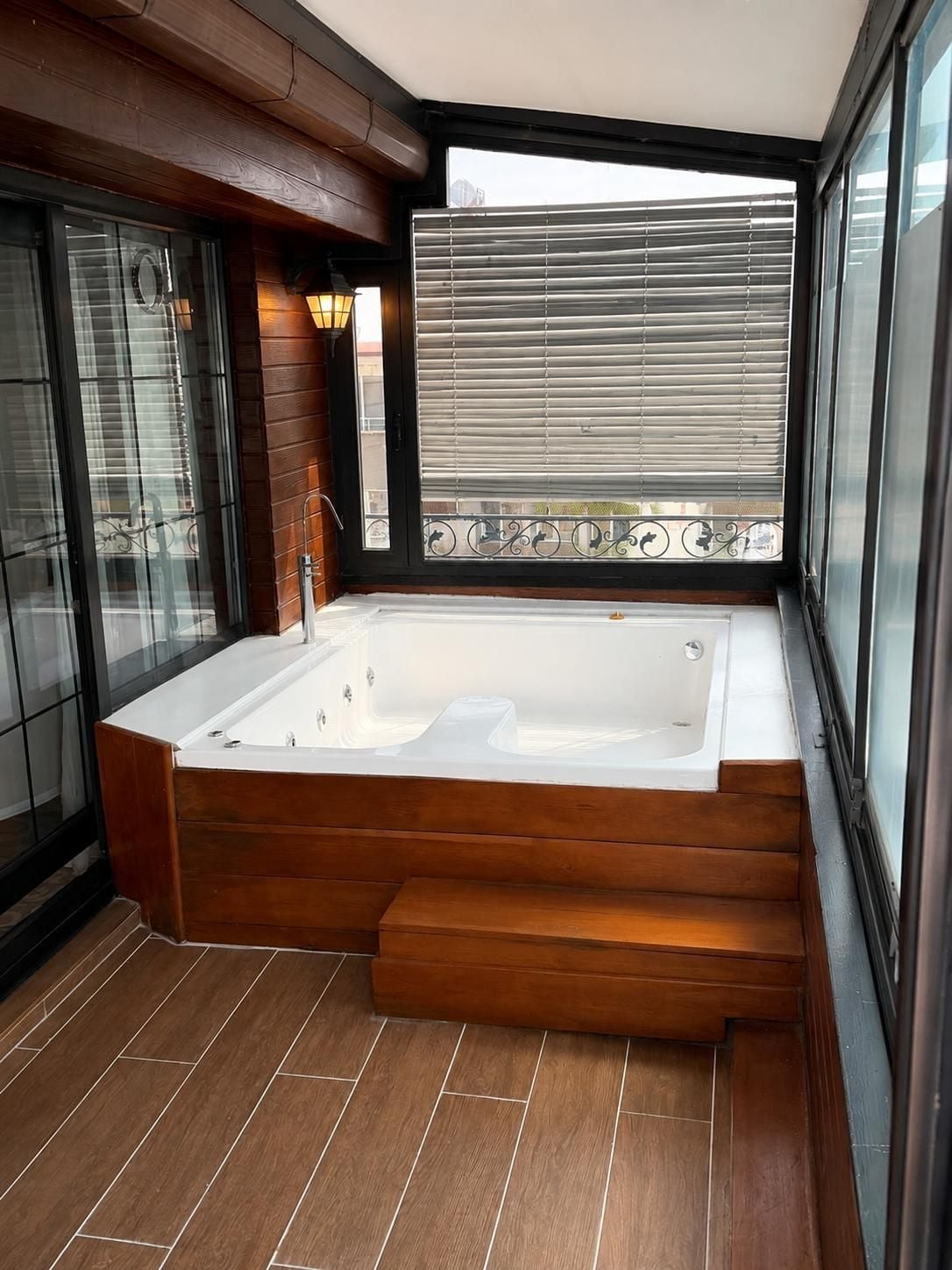 A modern bathroom with a freestanding bathtub, wooden flooring, and a large window with blinds.