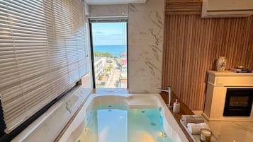 A modern bathroom with a glass-enclosed hot tub, wooden flooring, a marble wall, and a view of the sea.
