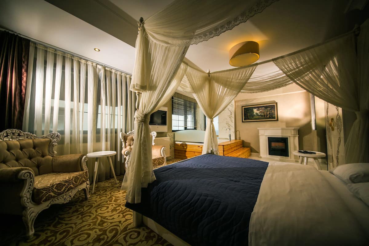 A bedroom with a four-poster bed, a sitting area with a chair and table, a fireplace, and a large window with curtains.