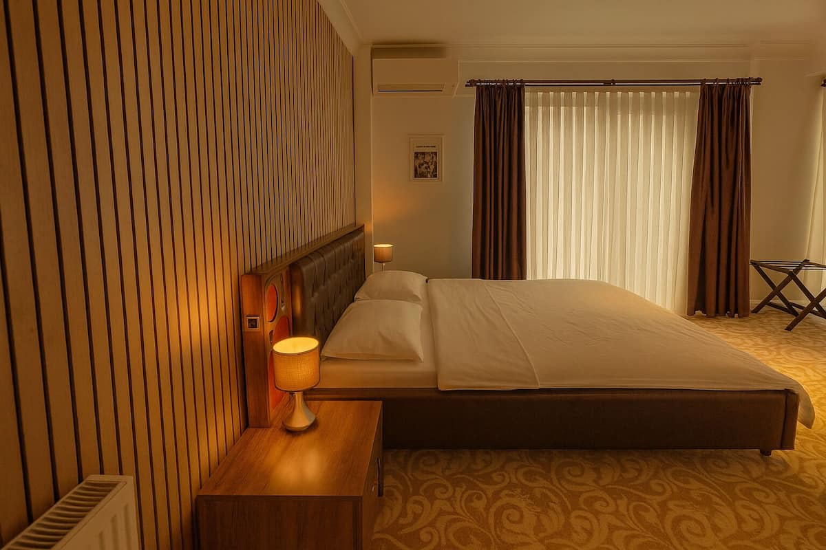 A hotel room with a bed, bedside table, lamp, curtains, and a small table with a chair.