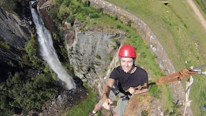 A person rappelling down a cliff with a rope, overlooking a waterfall and lush greenery.