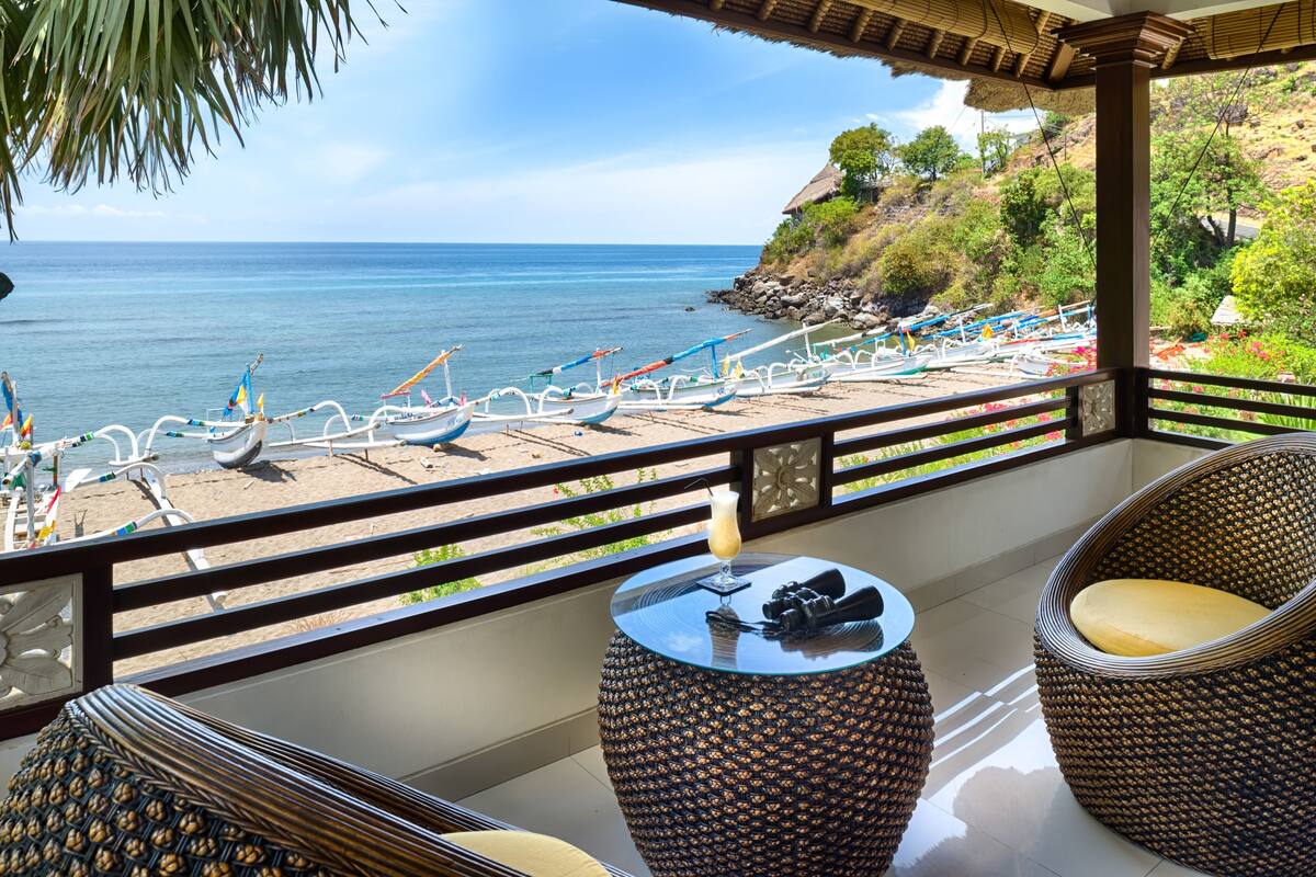 Un balcon agrémenté de meubles en osier, d’une table en verre et offrant une vue sur la plage, avec des bateaux et un bâtiment à toit de chaume.