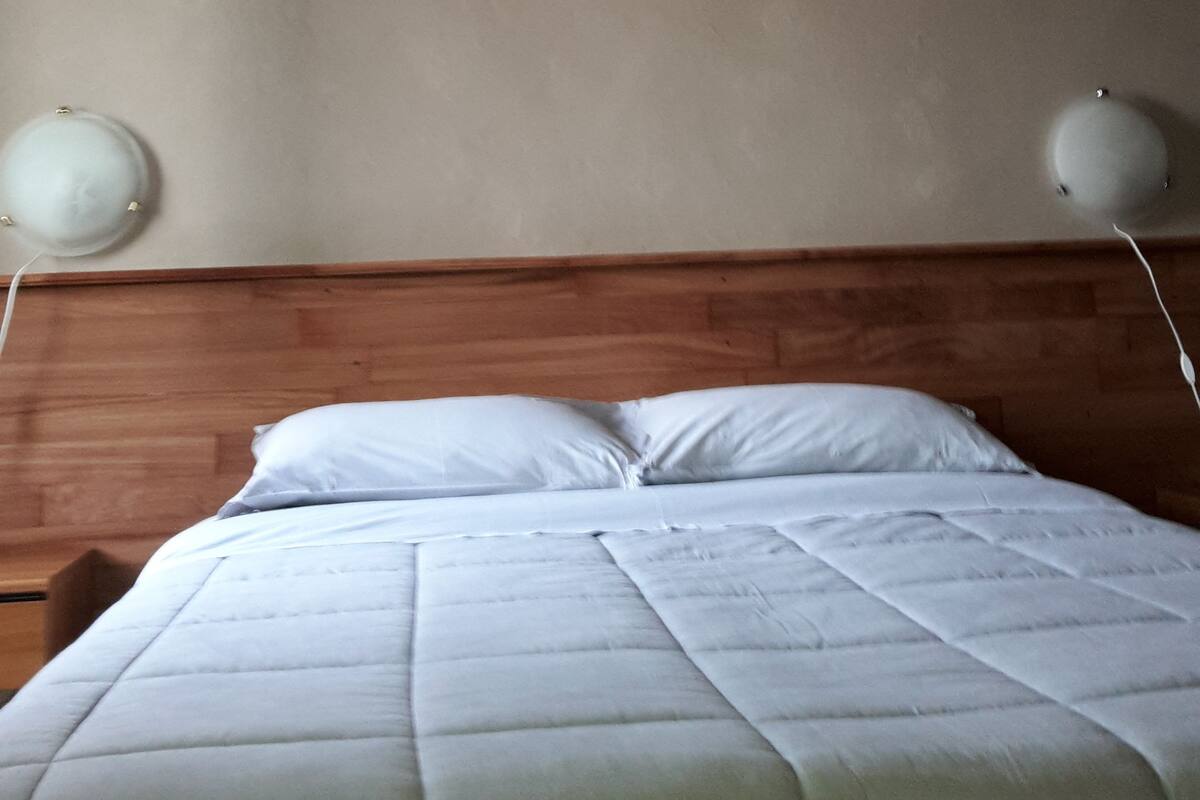 Een bed met een houten hoofdbord, een effen wand en twee wandlampen.