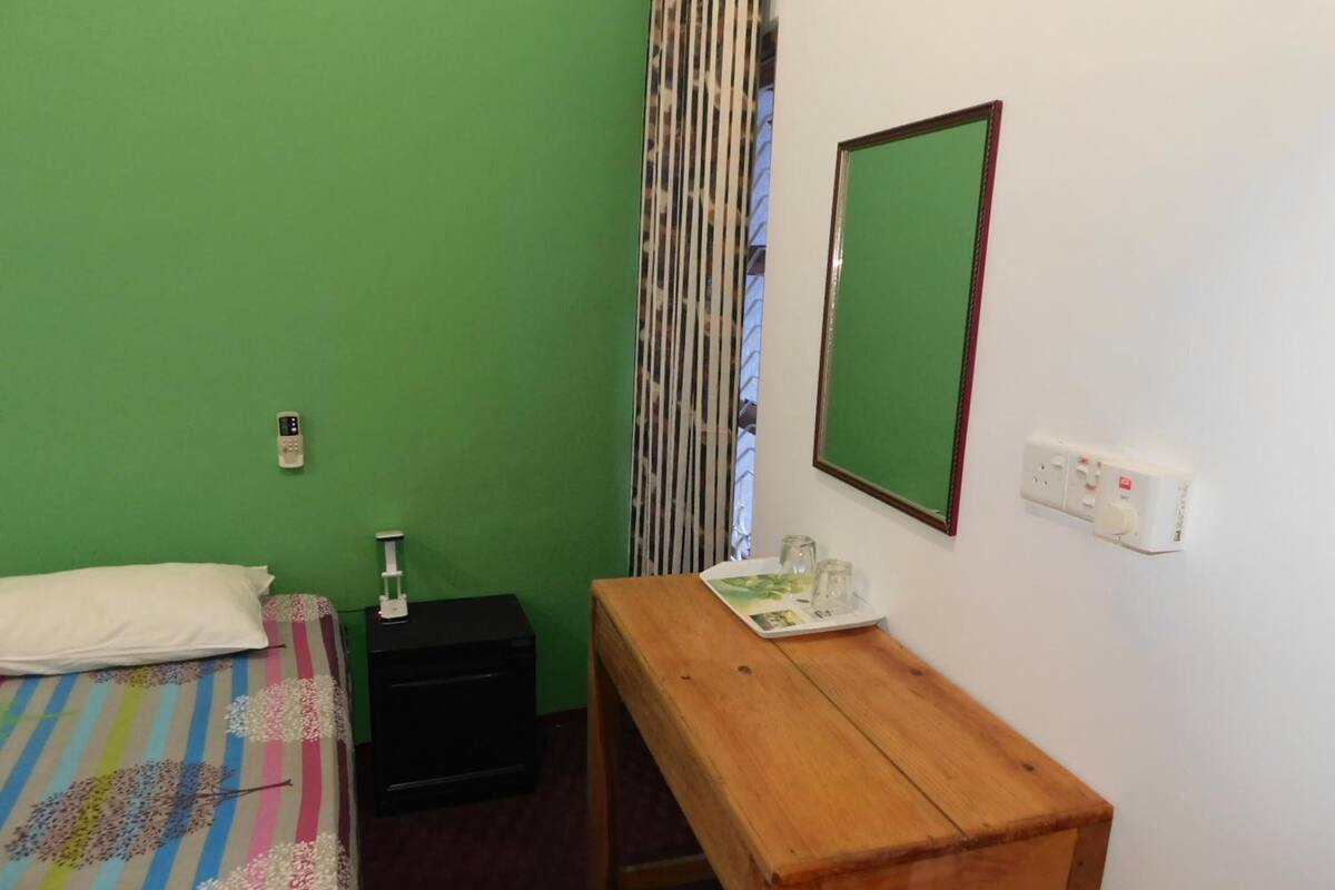 Quarto pequeno e simples, com parede verde, escrivaninha de madeira, cama com colcha listrada e colorida, mesa de cabeceira preta, espelho e janela com persianas verticais.
