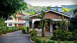 A two-story house with a stone facade, a balcony, and a garden with various plants and flowers. - Villa de Roses (Mahé Island)