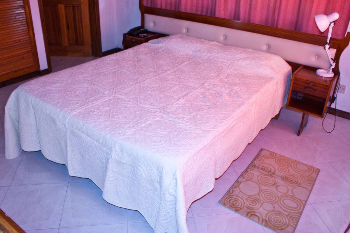Una cama bien hecha con una colcha blanca en una habitación con cabecera de madera y mesita de noche con lámpara.