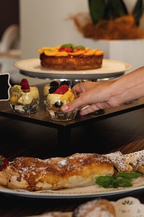 A dessert table with various pastries and a hand reaching for a dessert. - Hotel Europa (Malcesine)