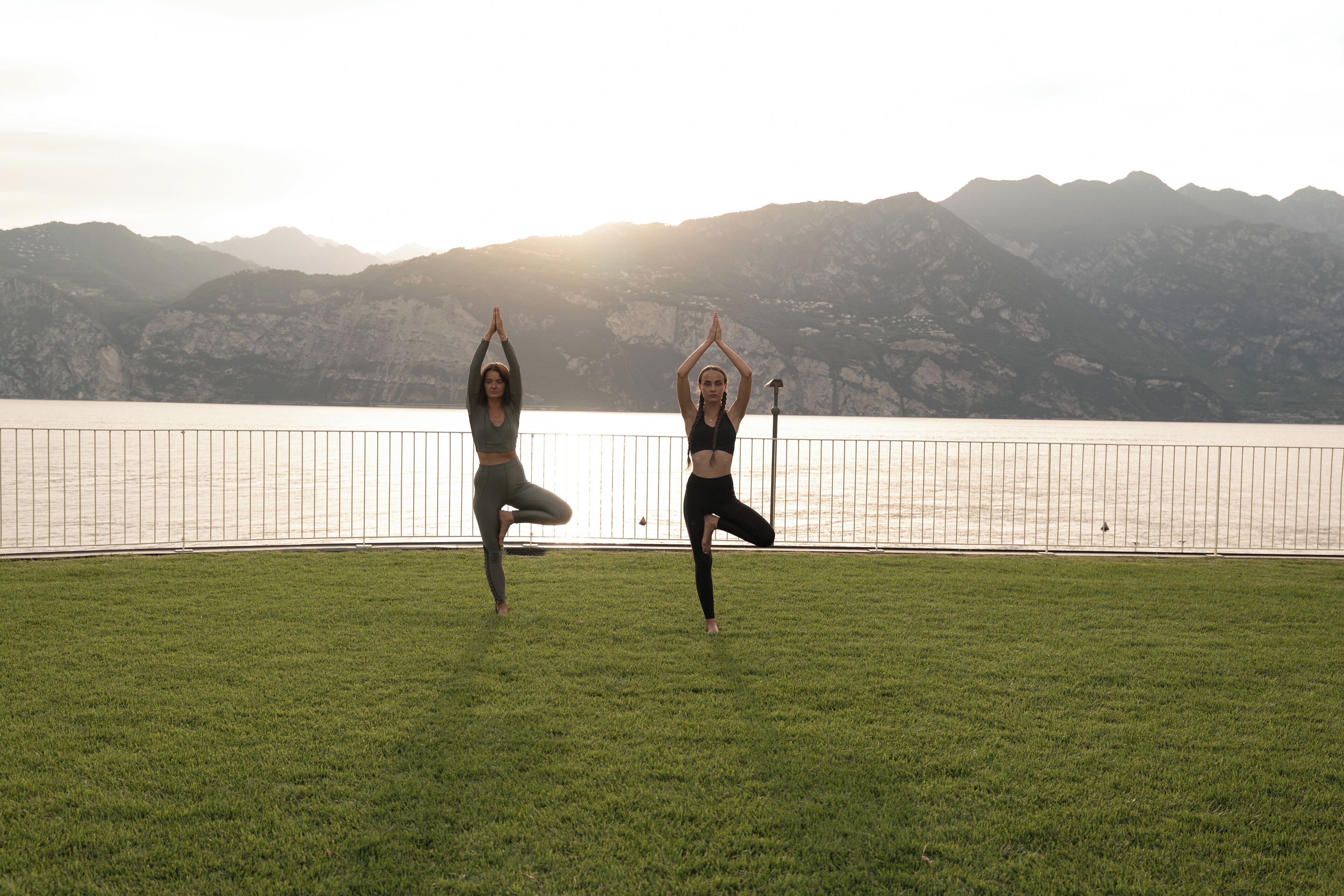 Two individuals performing yoga on a grassy area with a lake and mountains in the background.