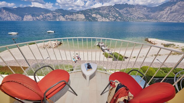 Een balkon met rode stoelen, een klein tafeltje met een boek, en een uitzicht op een meer met boten en bergen op de achtergrond.