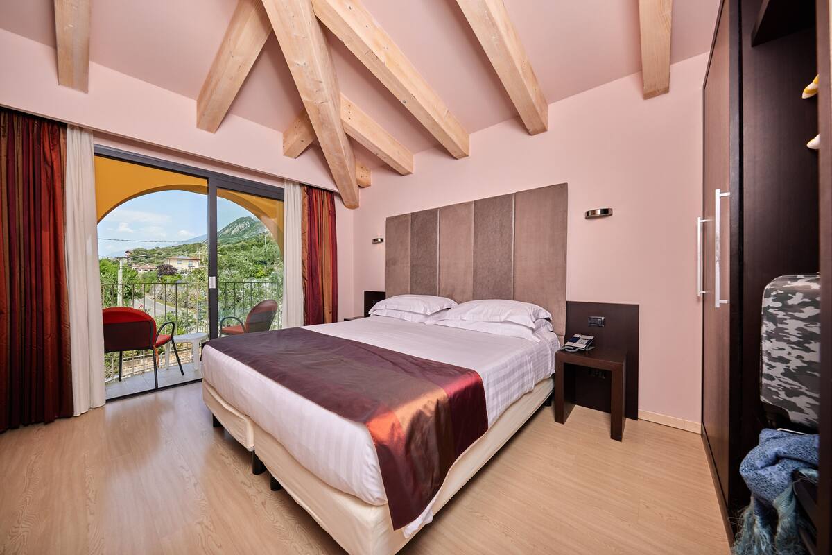 Une chambre moderne avec un grand lit, des poutres en bois au plafond, une porte-fenêtre coulissante donnant sur un balcon et une table de chevet avec une lampe.