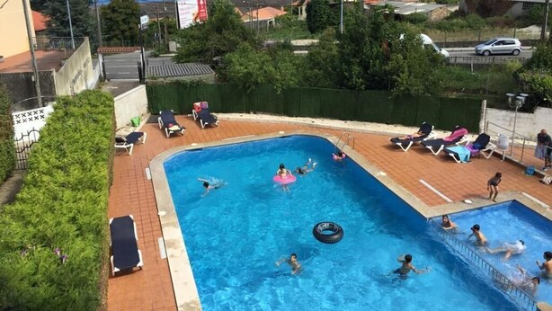 En swimmingpool med svømmende og afslappende mennesker, omgivet af grønt og udsigt til havet og bybillede.