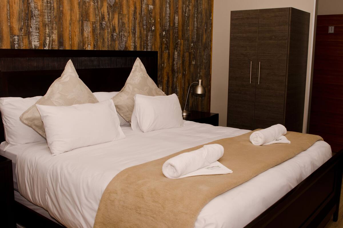 A neatly made bed with white linens and beige throw blankets, a wooden headboard, and a wooden wardrobe with two doors.