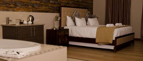 A hotel room with a large bed, a wooden headboard, a nightstand, a bedside table with a lamp, a bathroom with a bathtub, and a wooden floor.