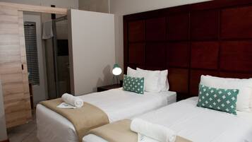 A hotel room with two beds, a wooden door, a glass shower, and a bedside lamp.