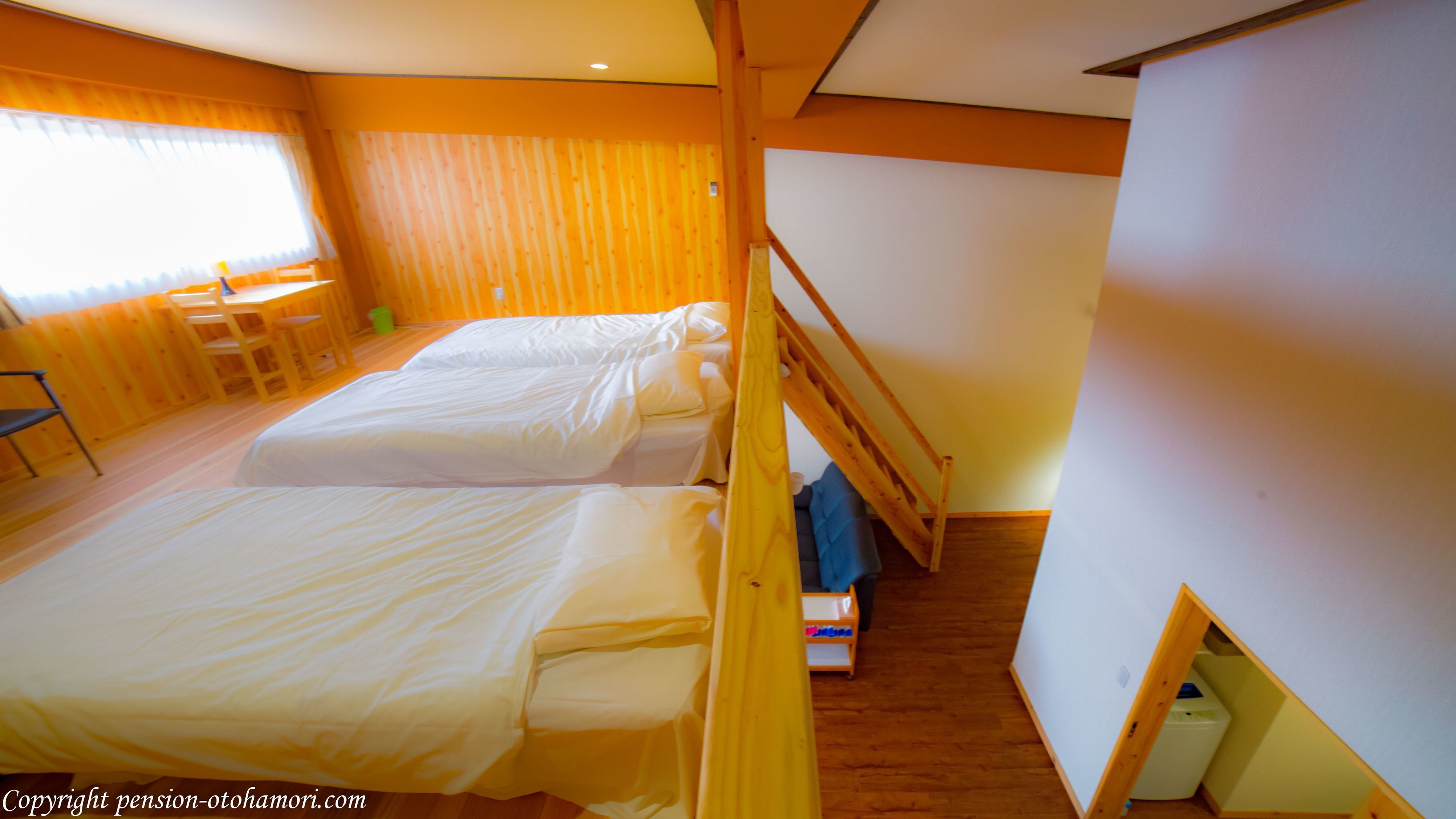 Ein Zimmer mit zwei Betten, Holzwänden, einem Fenster mit Vorhängen, einem kleinen Tisch und einem Stuhl.