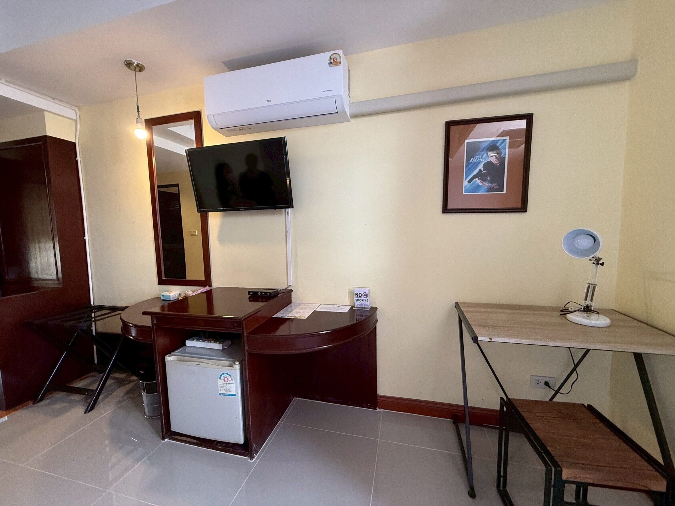Habitación de hotel con escritorio, televisión montada en la pared, un cuadro enmarco, refrigerador pequeño y lámpara de pie.