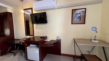 Habitación de hotel con escritorio, televisión montada en la pared, un cuadro enmarco, refrigerador pequeño y lámpara de pie.