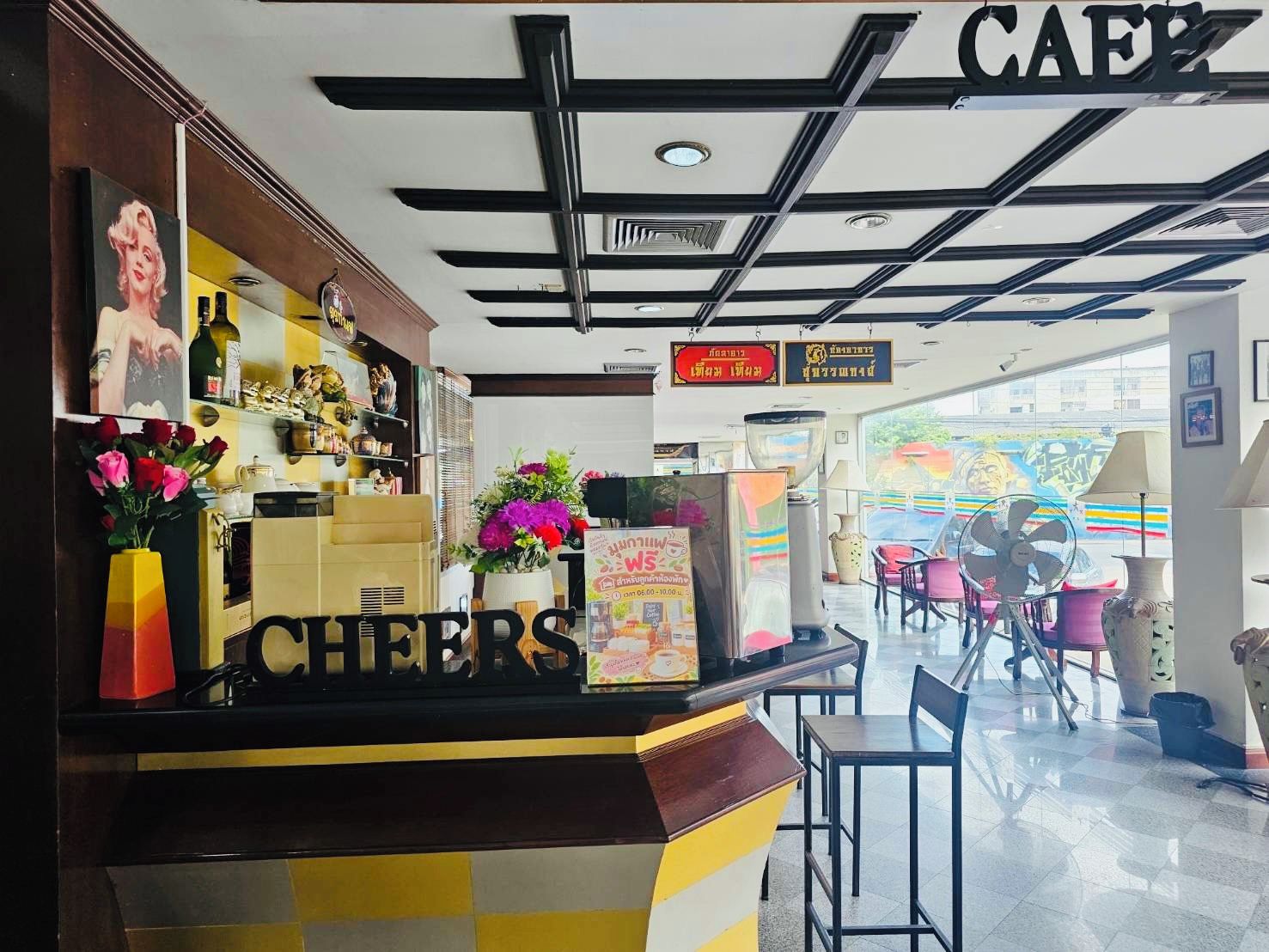 A cafe with a counter displaying 'CHEERS' and a 'CAFE' sign above. There are chairs and tables in the seating area.