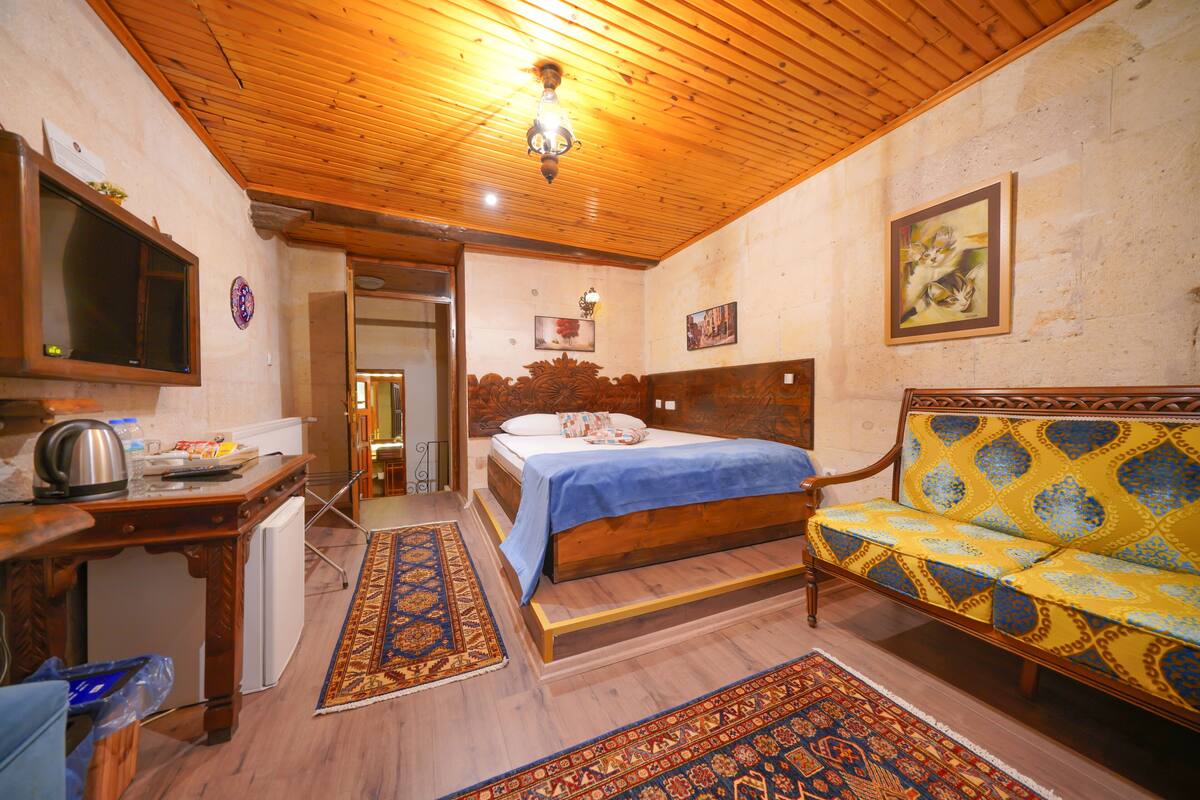 Une chambre avec un plafond en bois, un lit recouvert d’une courtepointe bleue, un banc en bois, un canapé agrémenté d’un coussin à motifs et un téléviseur fixé au mur.