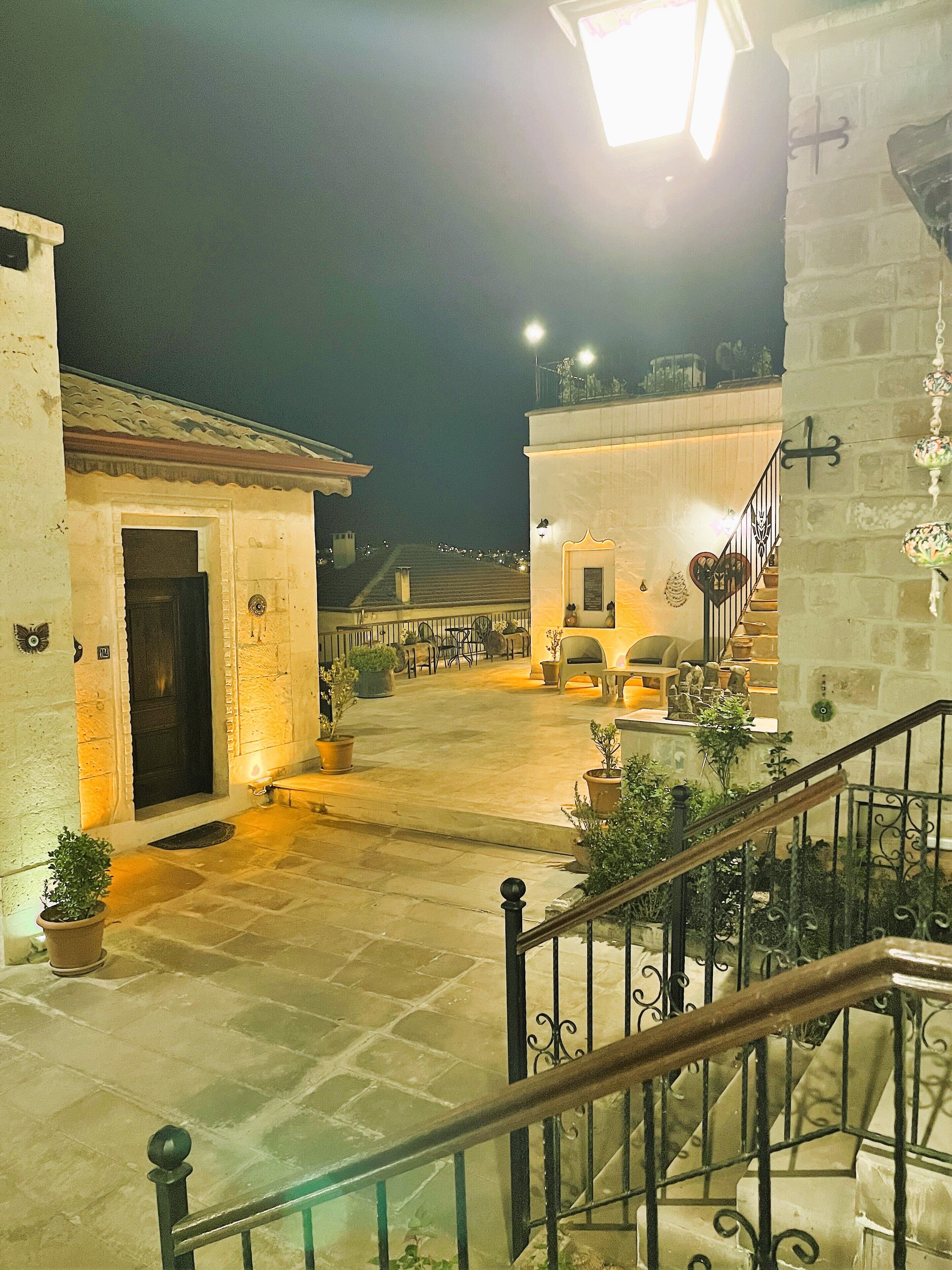 Un patio con una escalera que conduce a plantas superiores, macetas con plantas y una entrada bien iluminada.