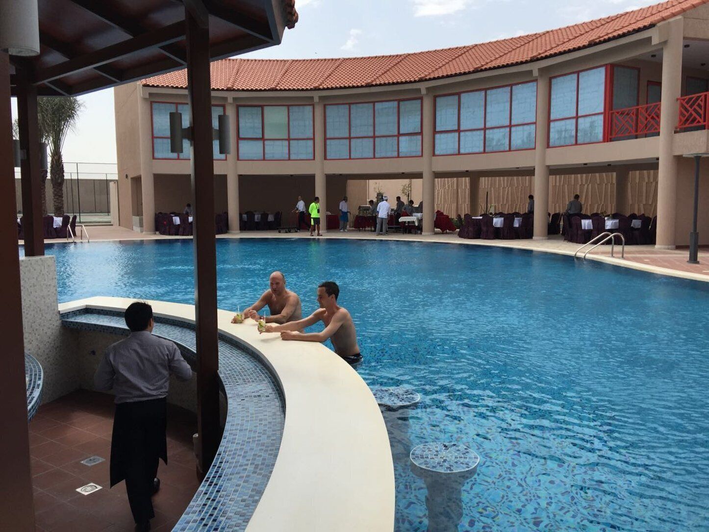 Une piscine à bord incurvé, un espace couvert avec des tables et des chaises, et un bâtiment avec de grandes fenêtres et un toit en tuiles en arrière-plan.