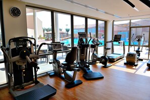A gym with various exercise machines, large windows, and a view of a swimming pool. - Al Reem Village Hotel (Al Jubail)
