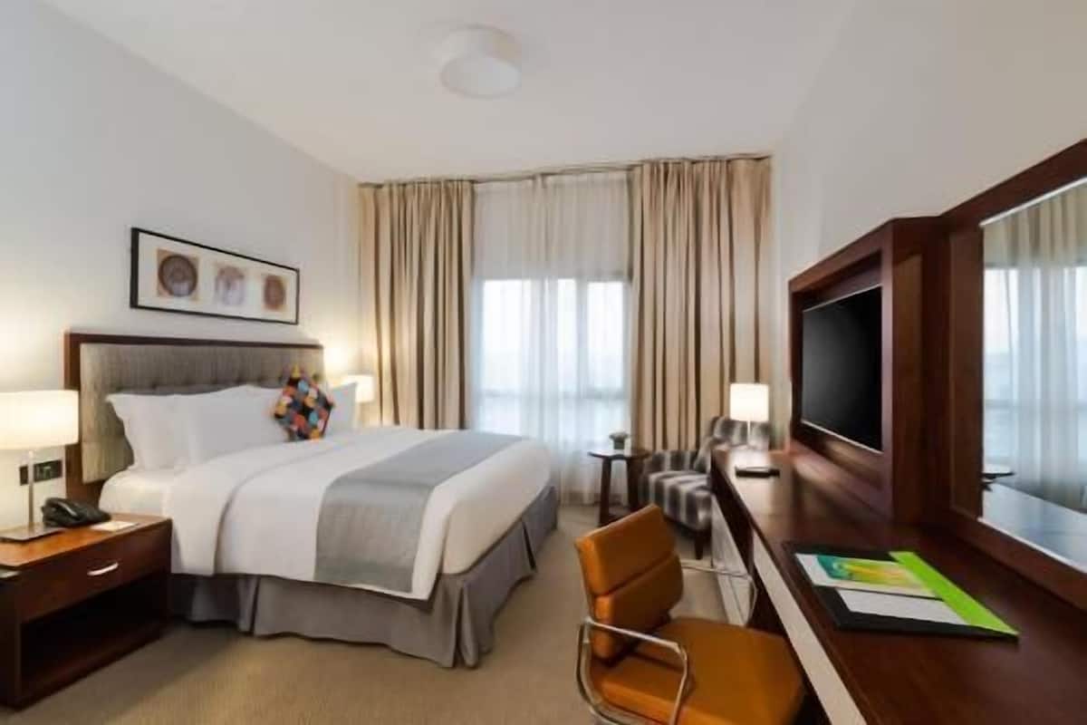 Ett hotellrum med en säng, ett skrivbord med en stol, en tv och ett fönster med gardiner.