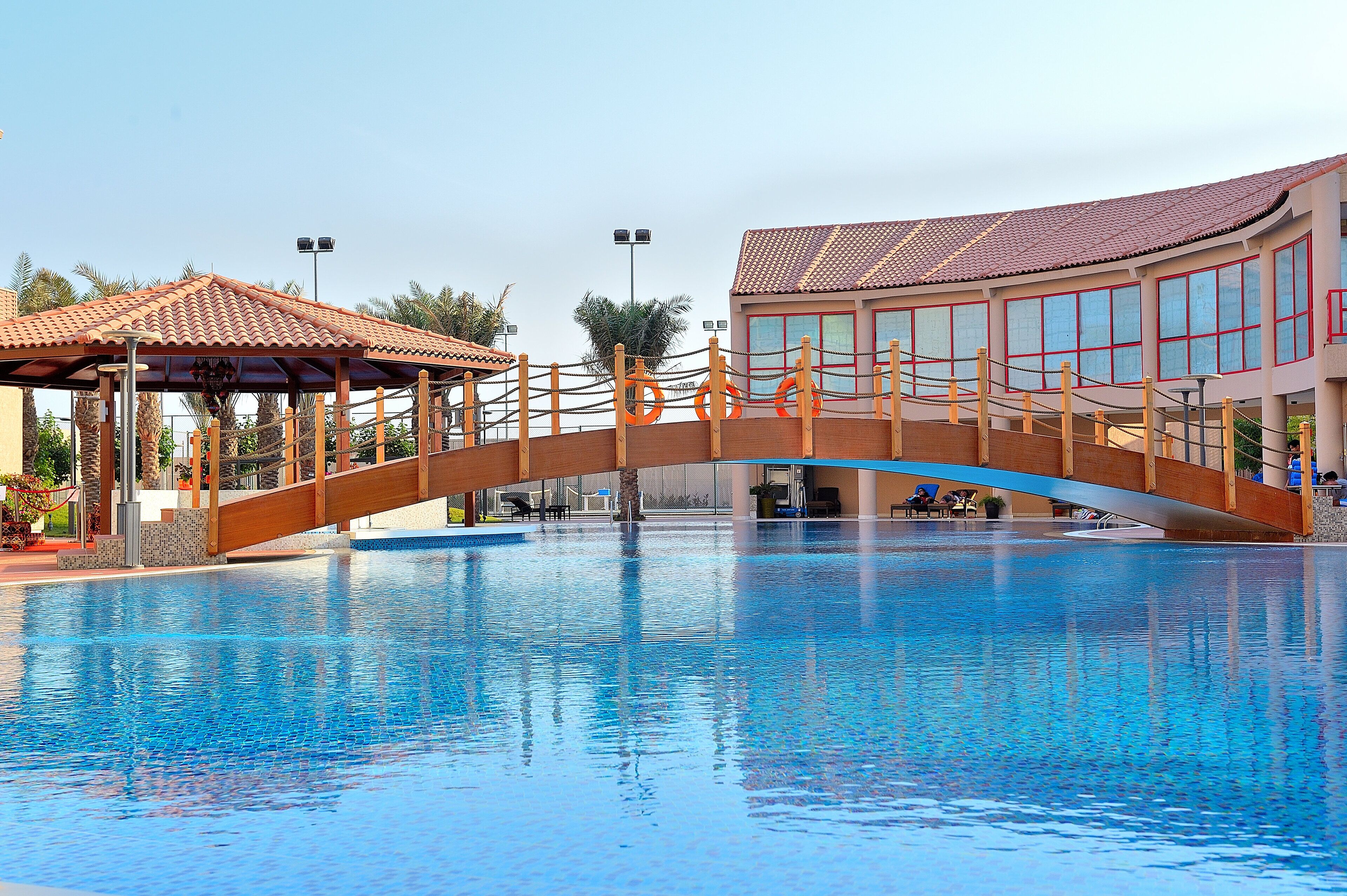 A swimming pool with a wooden bridge, a gazebo, and a building with large windows.